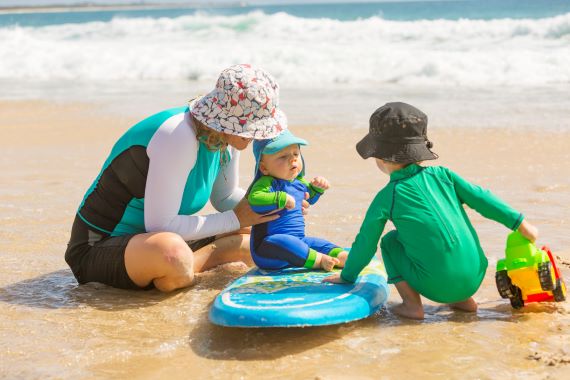 mother, baby and toddler on a beach wearing hats and rashguards