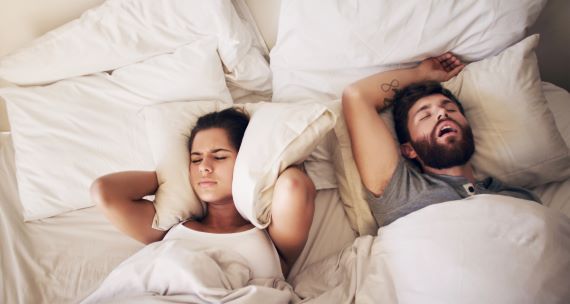 woman holding a pillow over her ears while her partner snores in bed next to her