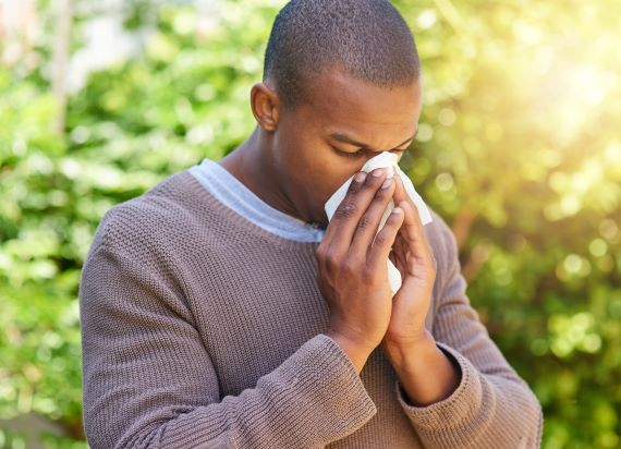 man blowing his nose into a tissue while standing in front of a tree on a sunny day