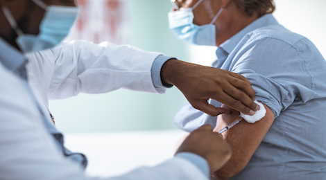 A doctor wearing a white coat and a blue mask injecting a shot into a man's left bicep