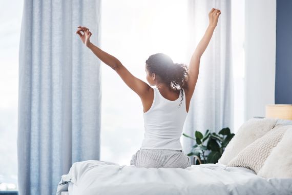 woman sitting on a bed stretching her arms overhead as the sun shines in the window