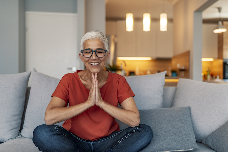 A woman meditates indoors on her couch.